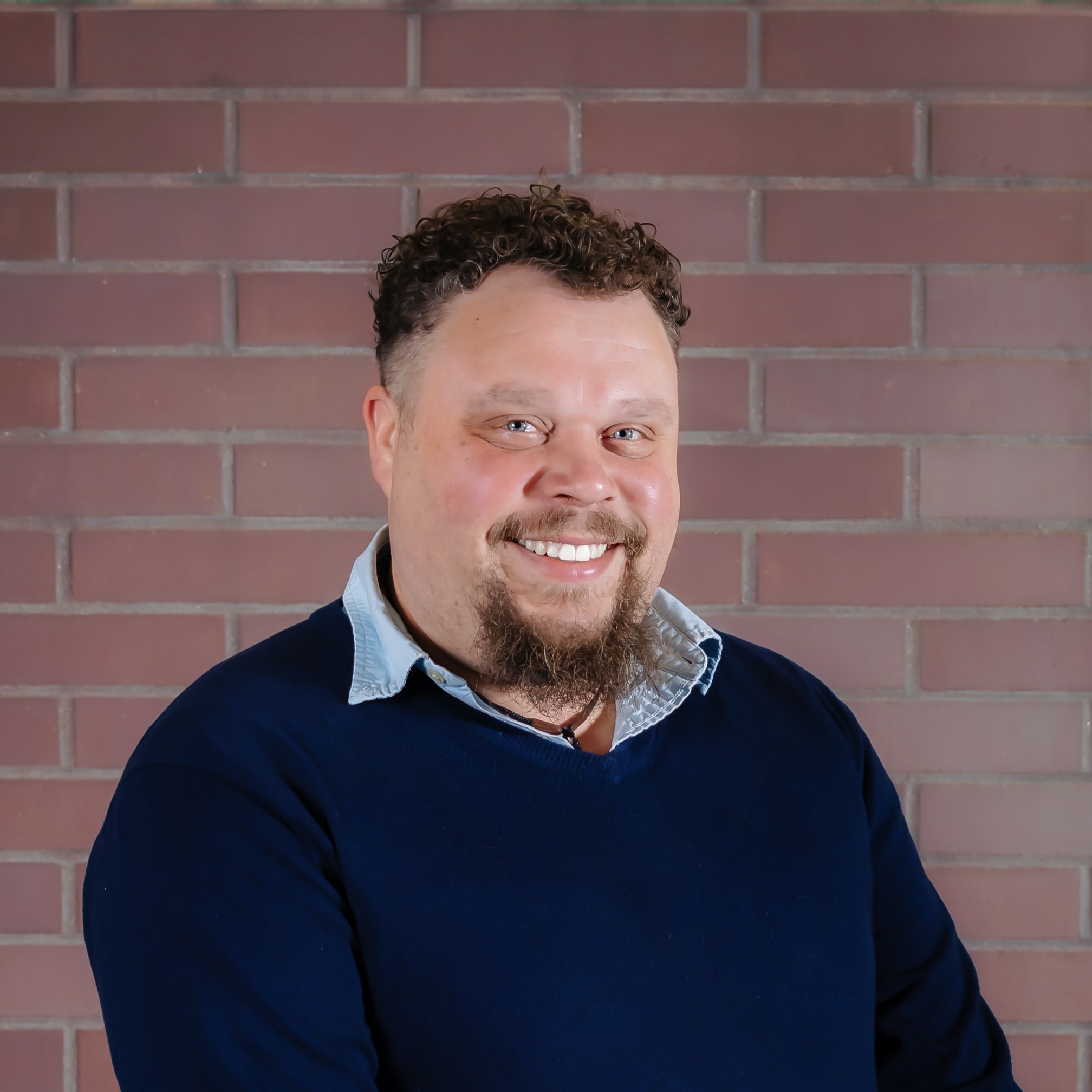 A headshot photo of a man with curly hair, a blue shirt and sweater, standing in front of a brick wall. The man is Jacob Campbell.